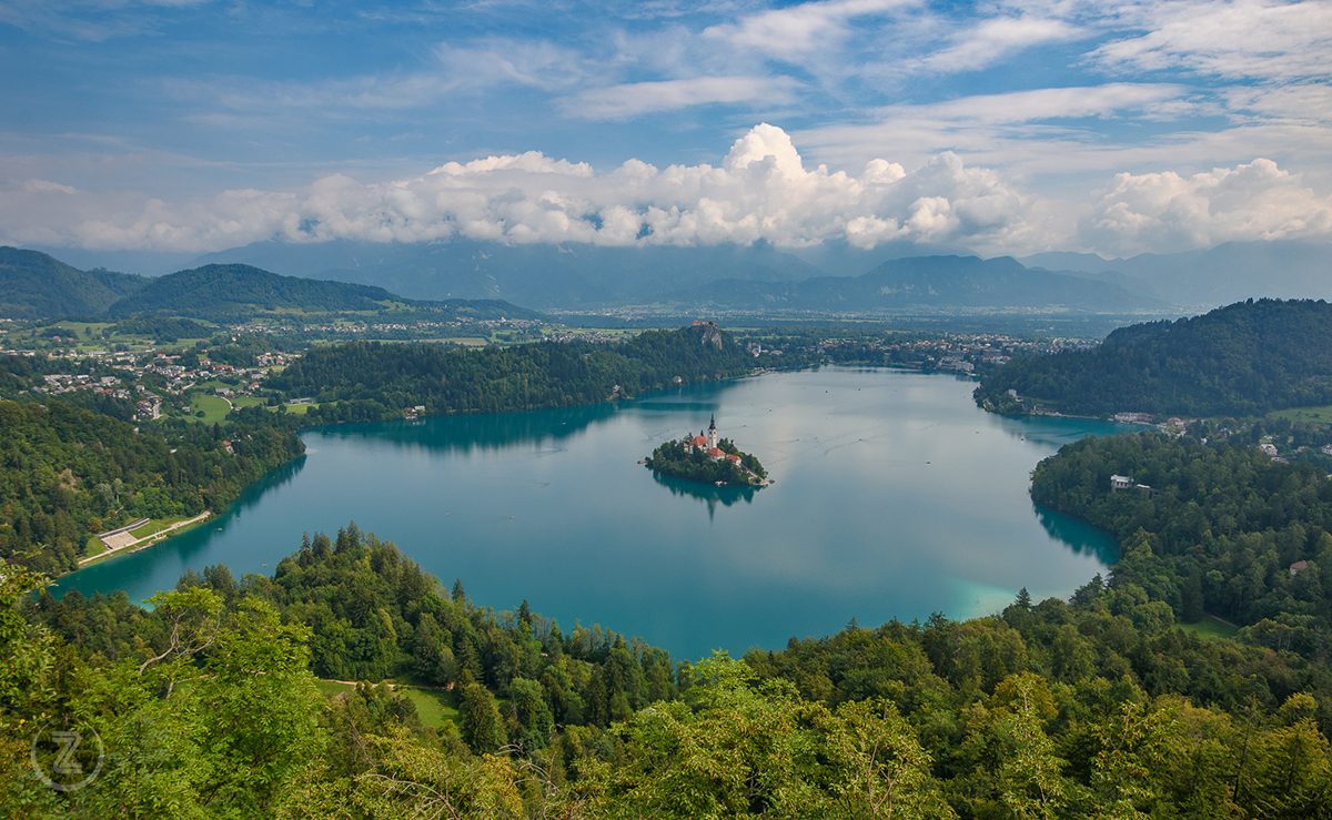 Panoramski pogled na Blejsko jezero z otokom in cerkvijo, Blejskim gradom ter gorami v ozadju.
