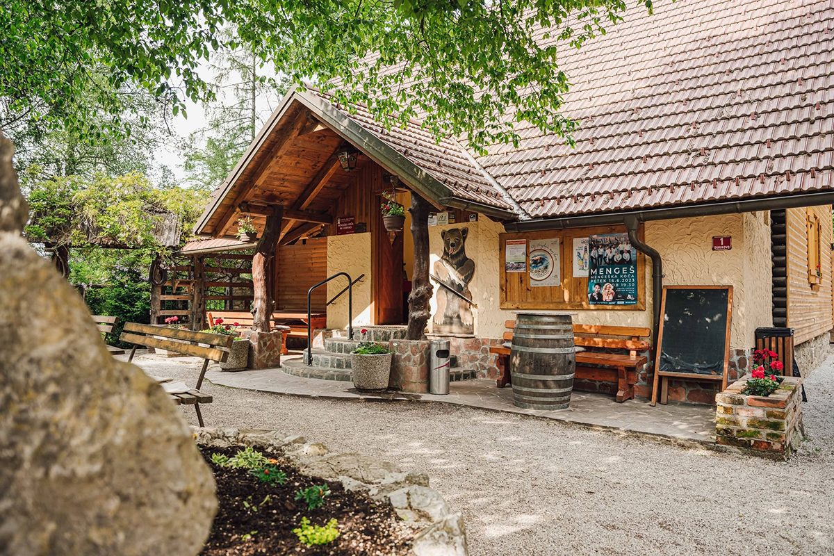 Vhod v Mengeško kočo na Gobavici z lesenim nadstreškom, klopmi in cvetjem v senci dreves.
